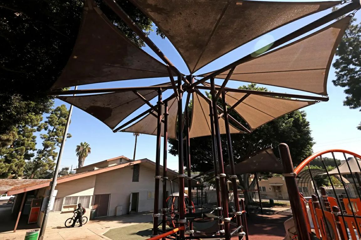 Large triangular fabric canopies shade playground equipment at the Hoover Recreation Center in Los Angeles. A bicyclist walks beside a low building in the background.