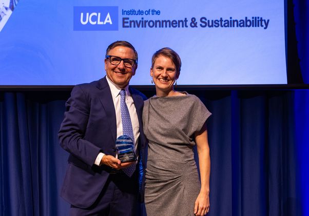 Tony Pritzker (left), in a navy suit, and Zuzana Buřivalová, in a gray dress, smile while holding an award onstage beneath a UCLA Institute of the Environment & Sustainability sign.