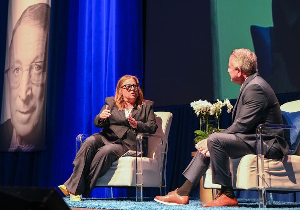 Cori Close gestures while speaking with Brian Cornell onstage at Pauley Pavilion, with a large portrait of John Wooden visible behind them.