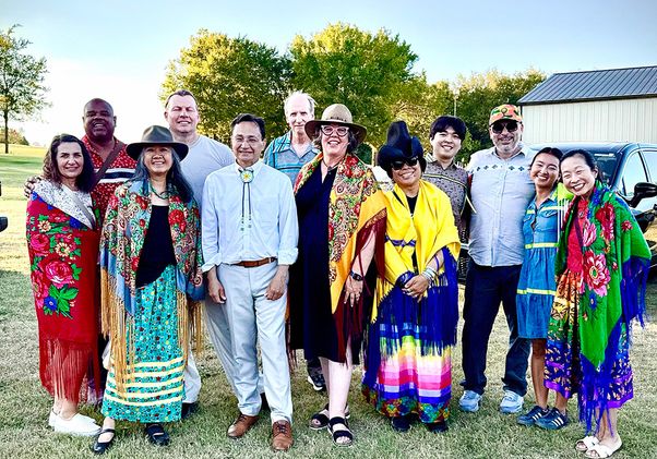 A diverse group of 12 adults pose together outdoors on a grassy field, wearing colorful traditional garments and shawls, with trees, a metal building and parked vehicles behind them.