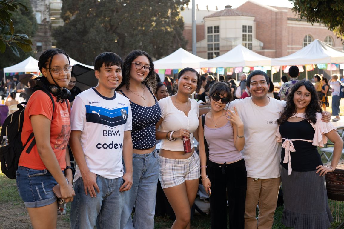 Seven attendees smile and pose together on a lawn at UCLA’s 2026 Latinx Welcome celebration, with white event tents, colorful decorations and other participants in the background.