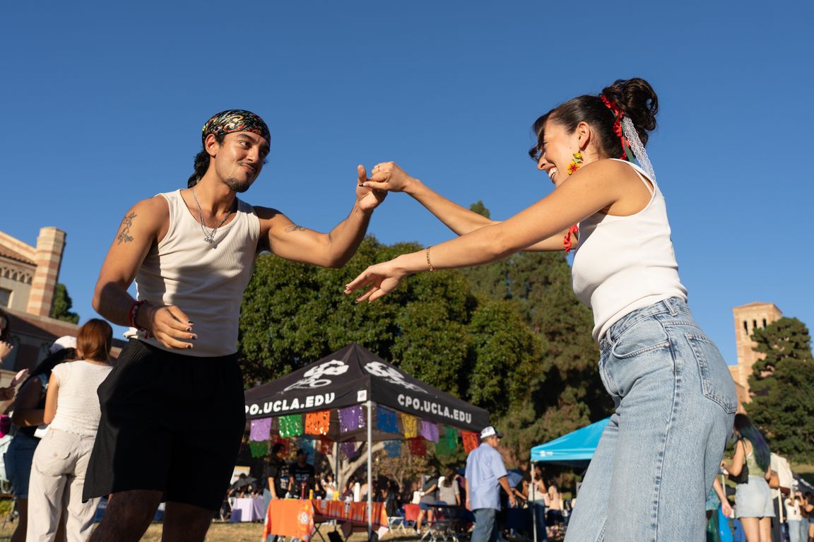 Two people smile and dance together outdoors at UCLA’s 2026 Latinx Welcome celebration, with colorful decorations, campus tents and other attendees in the background.