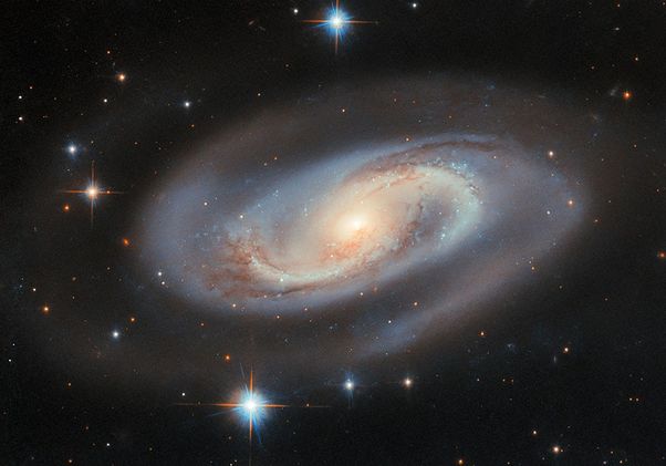 A glowing spiral galaxy with a bright yellow-white core, blue outer arms and dark dust lanes surrounded by stars against a black background.