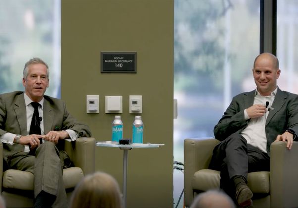 Kal Raustiala in suit and tie, left, and Evan Gershkovick in gray suit sit in armchairs during a talk, with water bottles on a small table between them and an audience in the foreground.