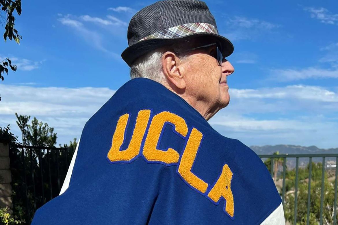 Andrew Ruane wearing a black fedora and sunglasses, draped in a blue and gold UCLA blanket, outdoors beneath a bright blue sky.