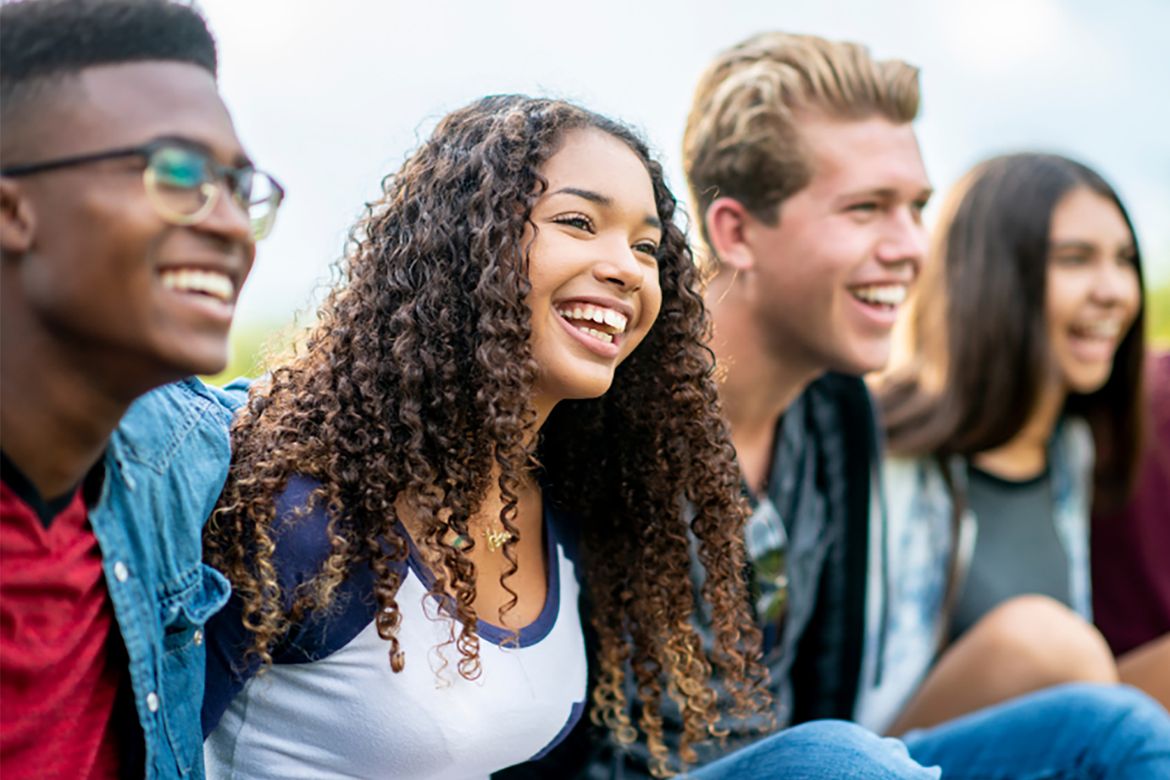 A diverse group of five young adults sitting outdoors, smiling and laughing together in a relaxed social setting.