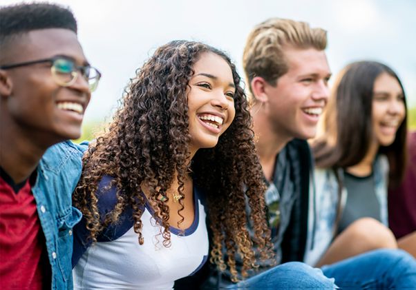 A diverse group of five young adults sitting outdoors, smiling and laughing together in a relaxed social setting.