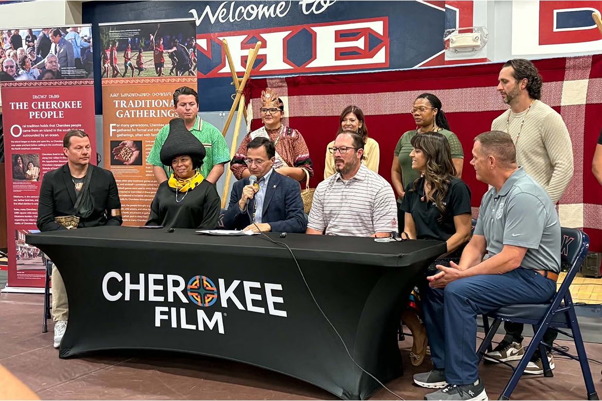 A diverse group gathers around a table labeled “Cherokee Film” during a panel discussion, with seated speakers and standing attendees in traditional and contemporary clothing.
