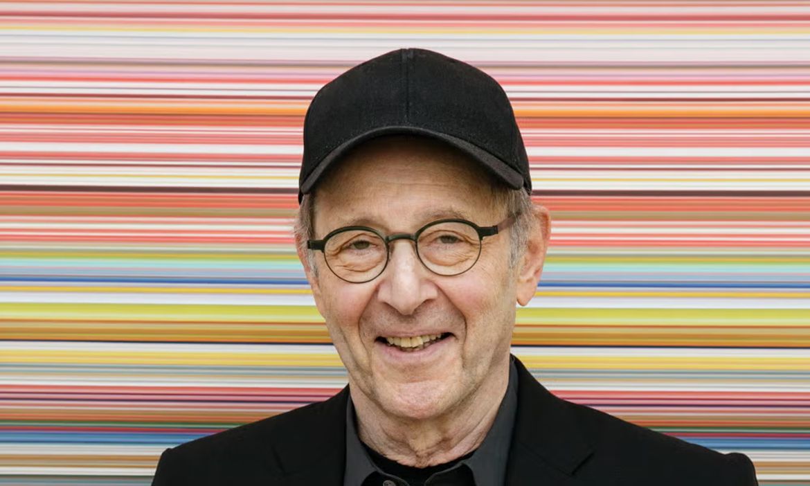 An older man in a black hat, smiling into the camera in front of a colorful striped background.