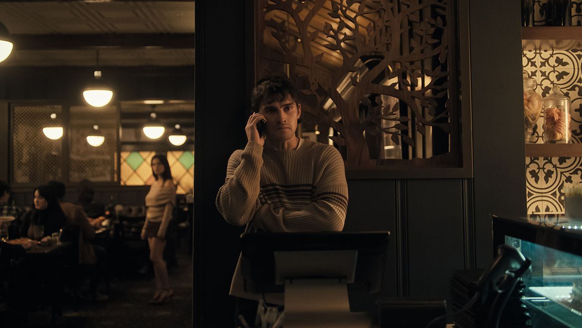 A young man in a striped sweater speaks on his phone behind a restaurant counter, surrounded by warm lighting, decorative wood panels and seated diners.