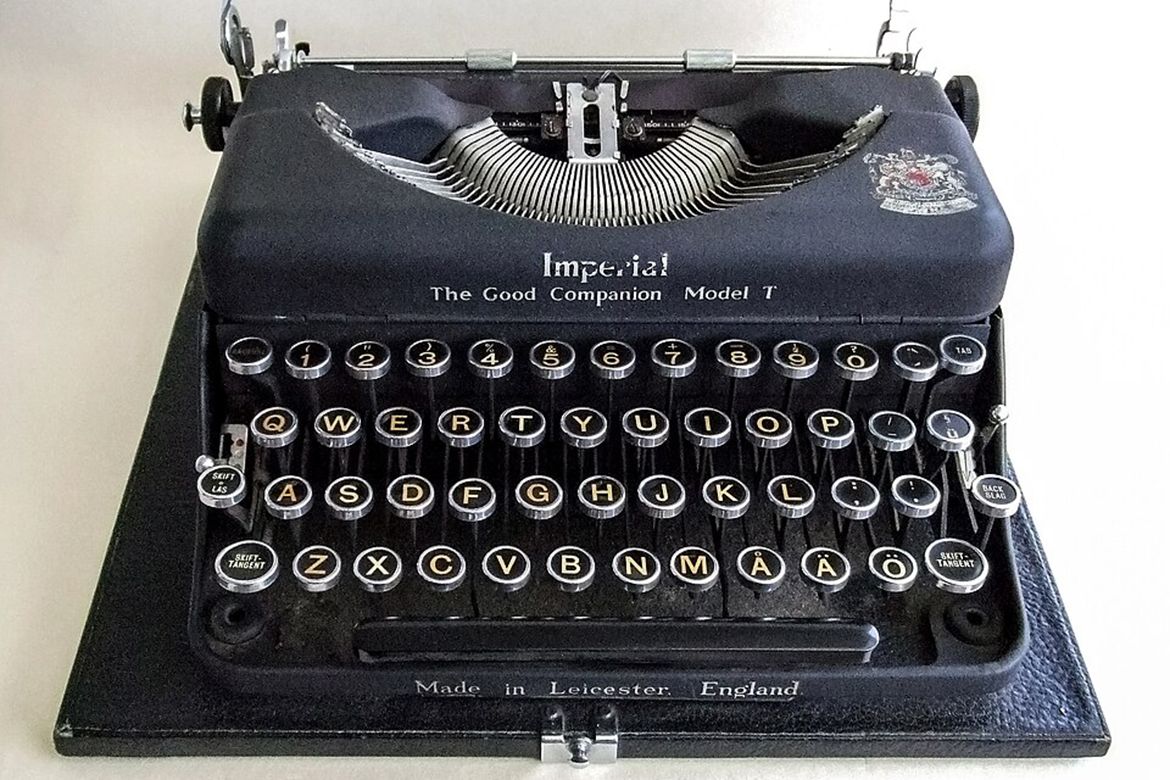 A black Imperial Good Companion Model T typewriter with round keys, exposed metal typebars and a textured base, labeled “Made in Leicester, England.”