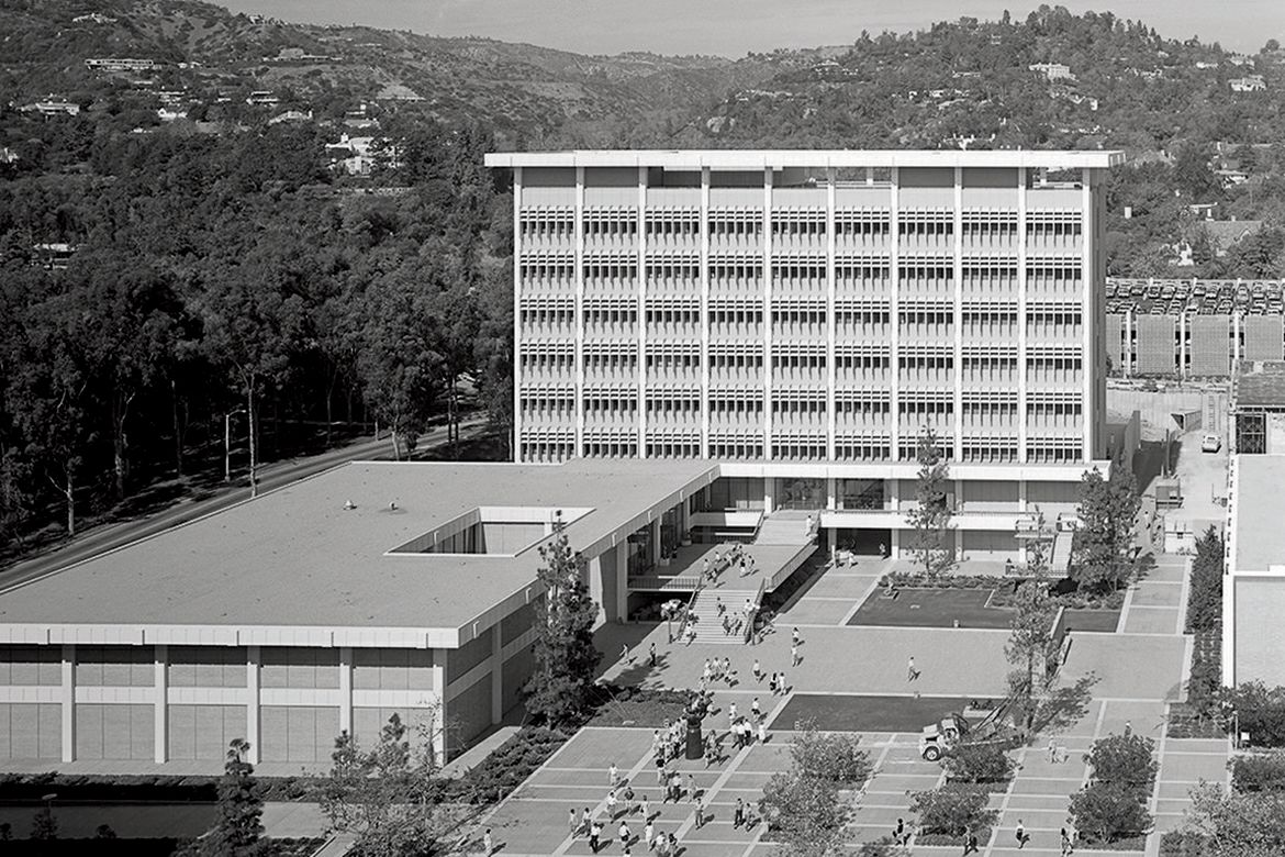 Black-and-white aerial view of a modern multi-story institutional building, adjacent low-rise structure, landscaped courtyard, paved walkways and surrounding wooded hills.