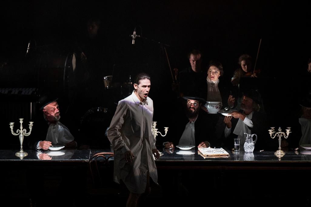 A theatrical scene shows a man in a white robe standing before a dark banquet table, surrounded by seated performers, musicians and tableware.