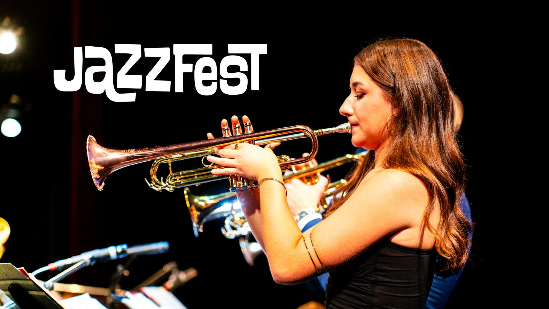 A young woman playing the trumpet during a live performance, spotlighted by a warm lighting. The text "Jazz Fest" is displayed on the top left side of the image.