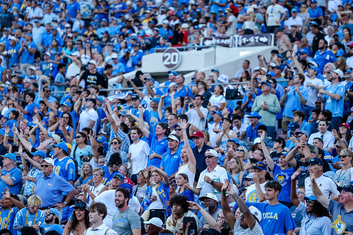 A packed stadium crowd of UCLA fans in blue and gold cheer, clap and raise their arms from the bleachers during a sporting event.
