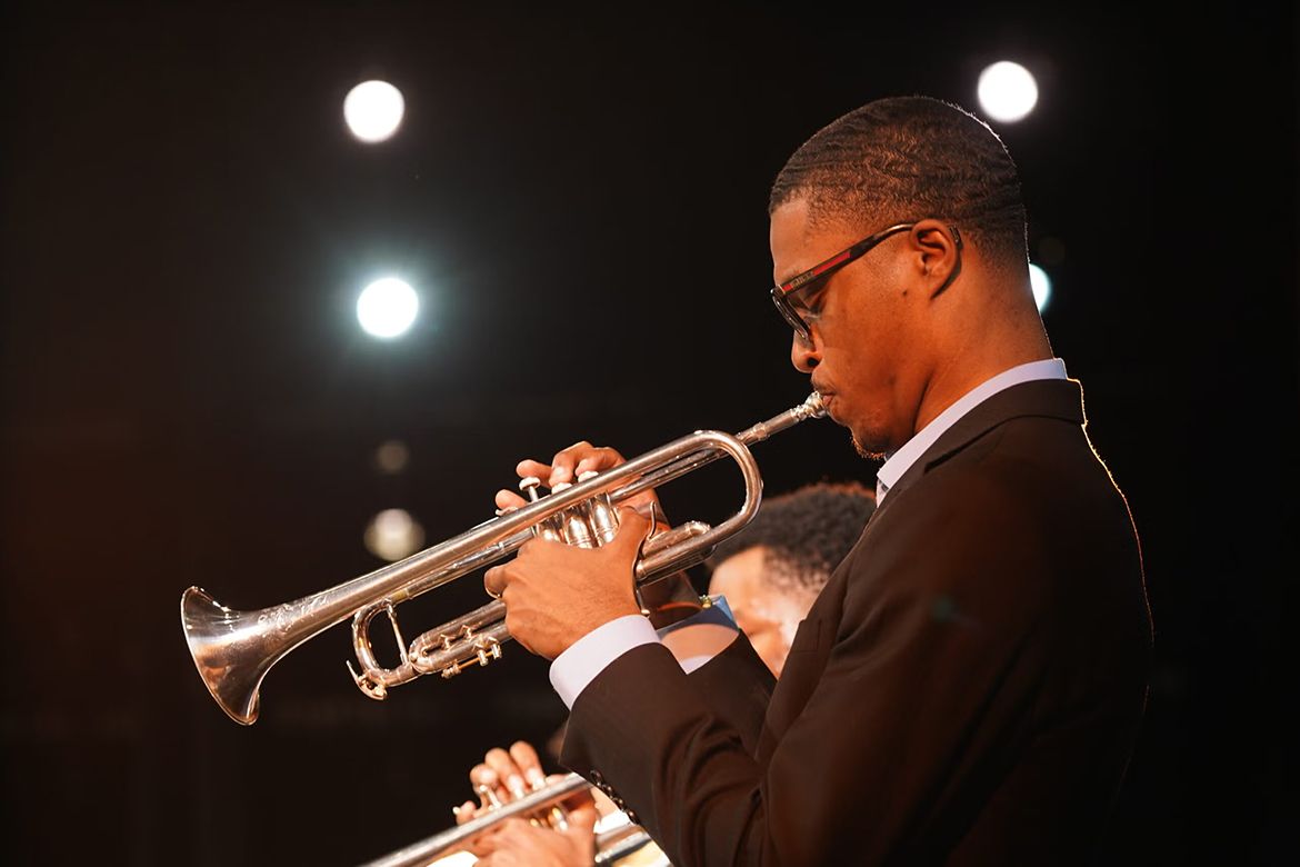 A musician wearing a black suit and glasses plays a silver trumpet onstage under bright lights, with other brass musicians partially visible behind him.