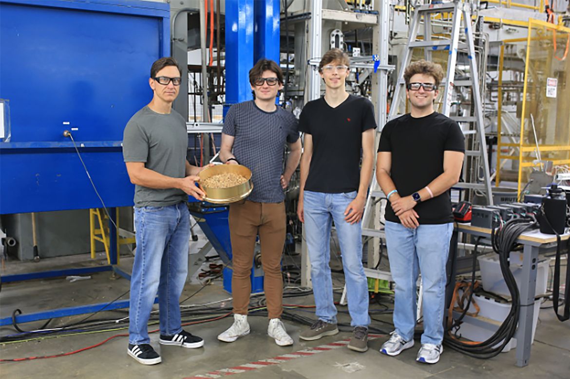 From left: Dante Simonetti, James Kasten, Alex Geldmeyer and Vagelis Christofides wear safety glasses while standing in an industrial workshop with machinery and equipment. Simonetti holds a metal bowl filled with small granular materials.