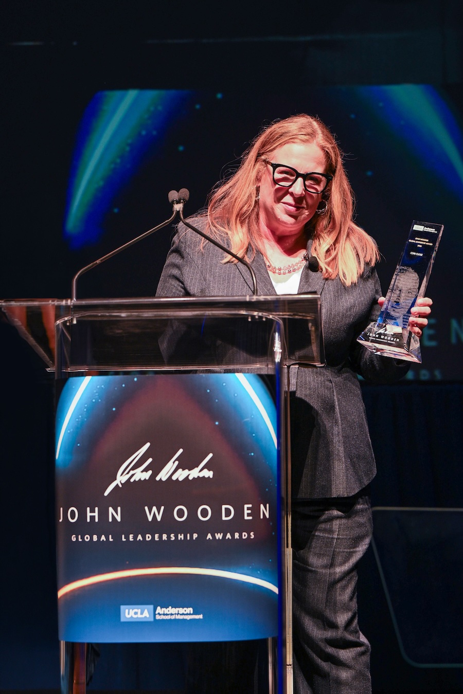 Cori Close smiles while holding a glass award behind a clear lectern bearing John Wooden’s signature.