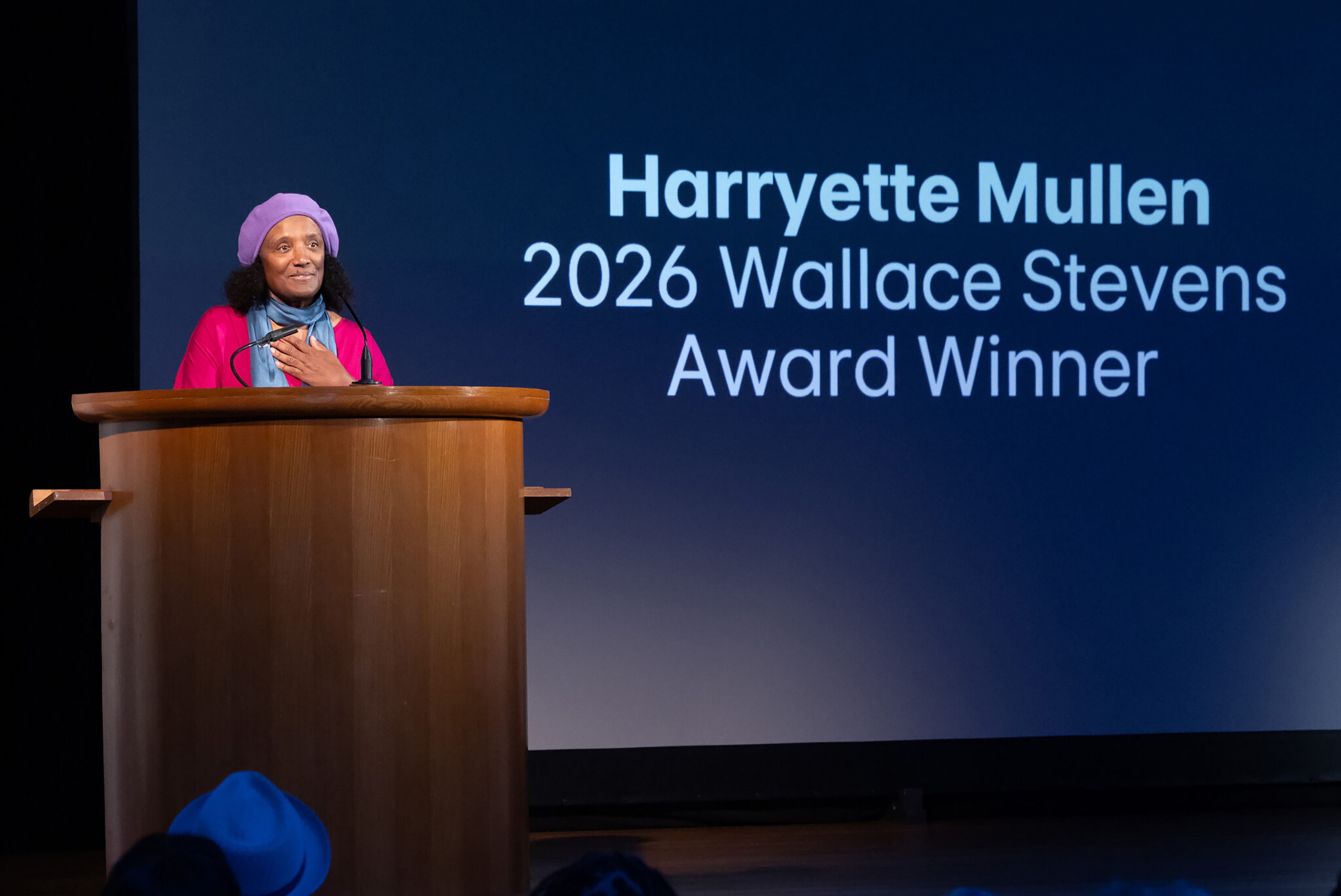 Harryette Mullen, wearing a purple beret and pink jacket, speaks at a wooden podium before a screen announcing her as the 2026 Wallace Stevens Award winner.