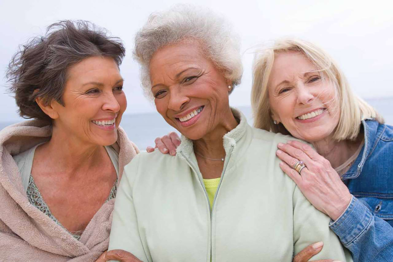 Three older women smiling and embracing outdoors, standing close together with their arms around one another against a bright, softly blurred background.