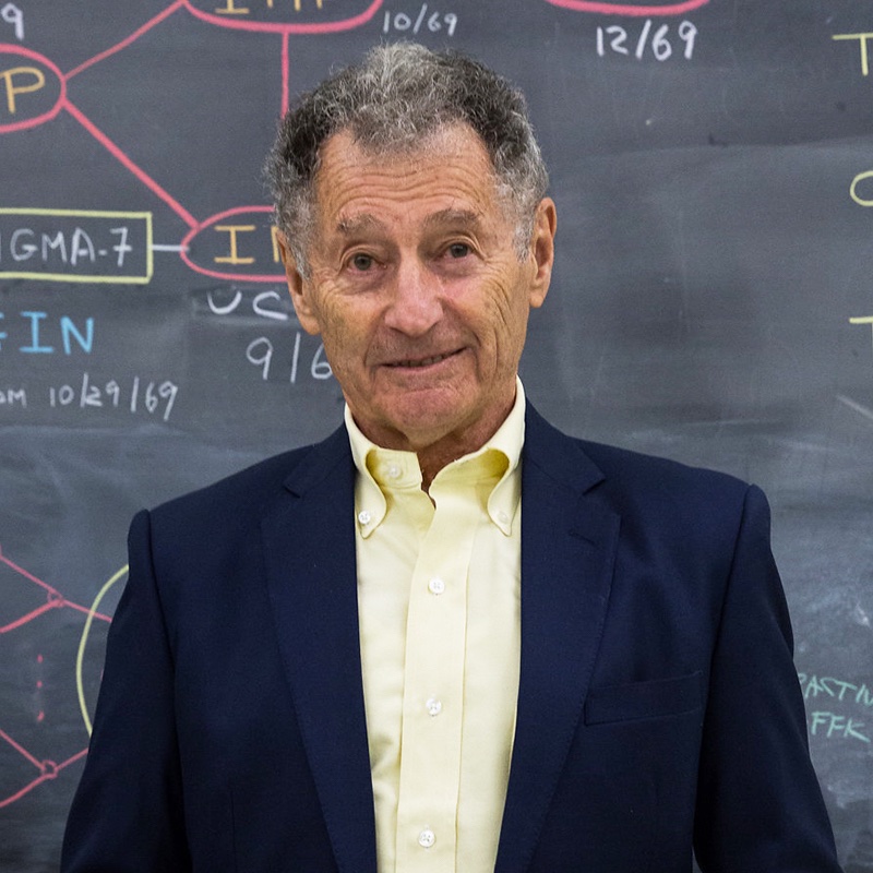 Leonard Kleinrock, wearing a navy blazer and pale yellow shirt, stands in front of a chalkboard covered with handwritten notes and diagrams.