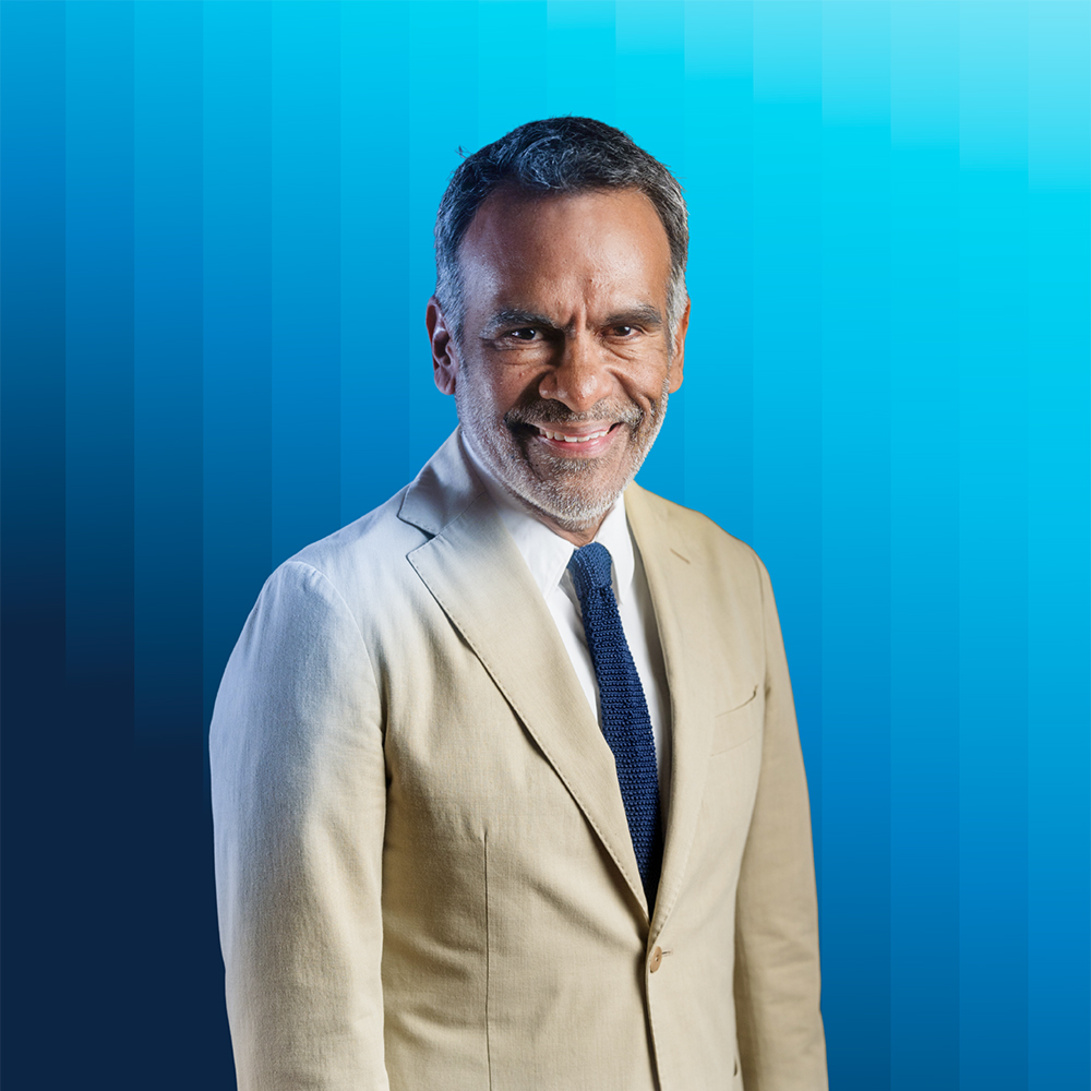 Portrait of Eric Avila in tan jacket and blue tie against a blue background.