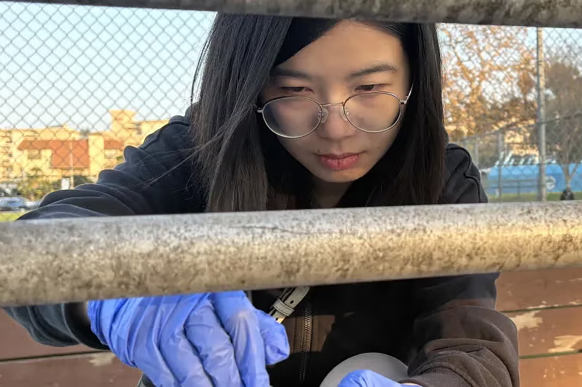 Yuan Yao wearing glasses, a black jacket and blue gloves leans over a metal railing, carefully examining an object outdoors.