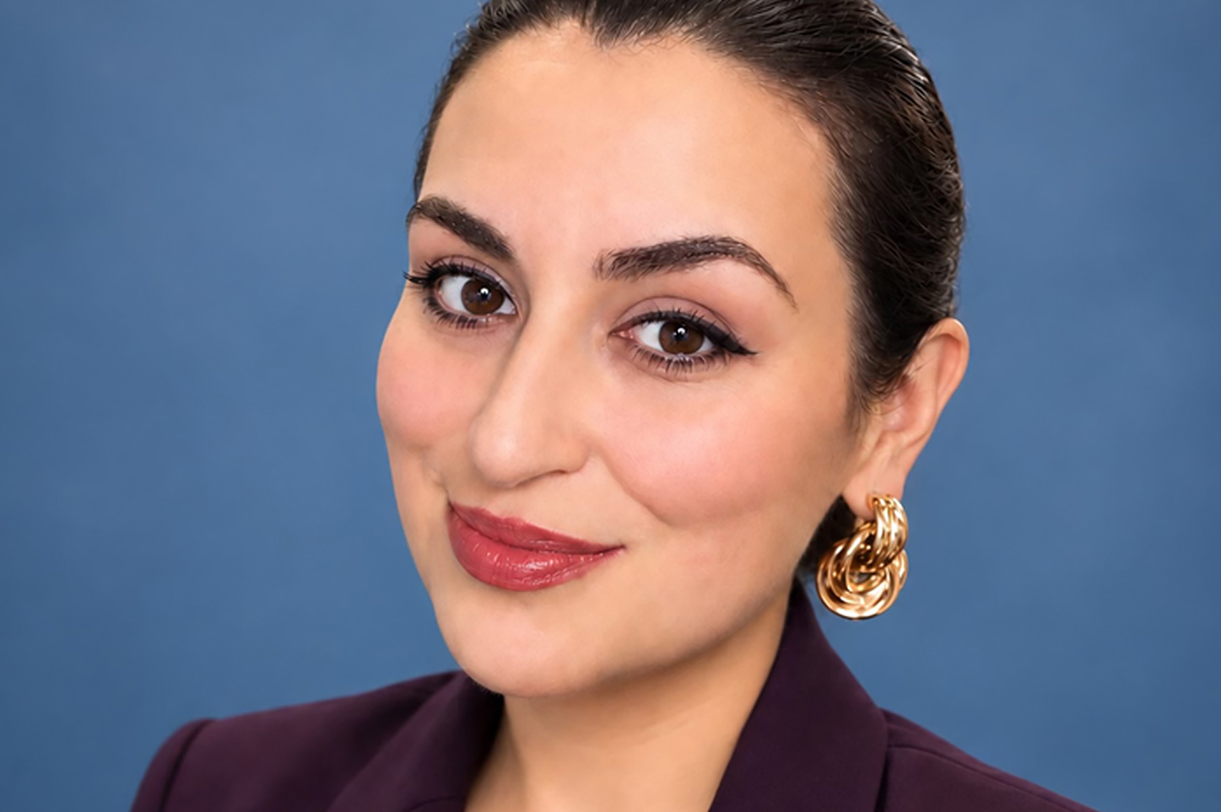  close  Close-up portait of Veronica Reynoso with hair pulled back, wearing gold statement earrings and a plum blazer against a blue background.