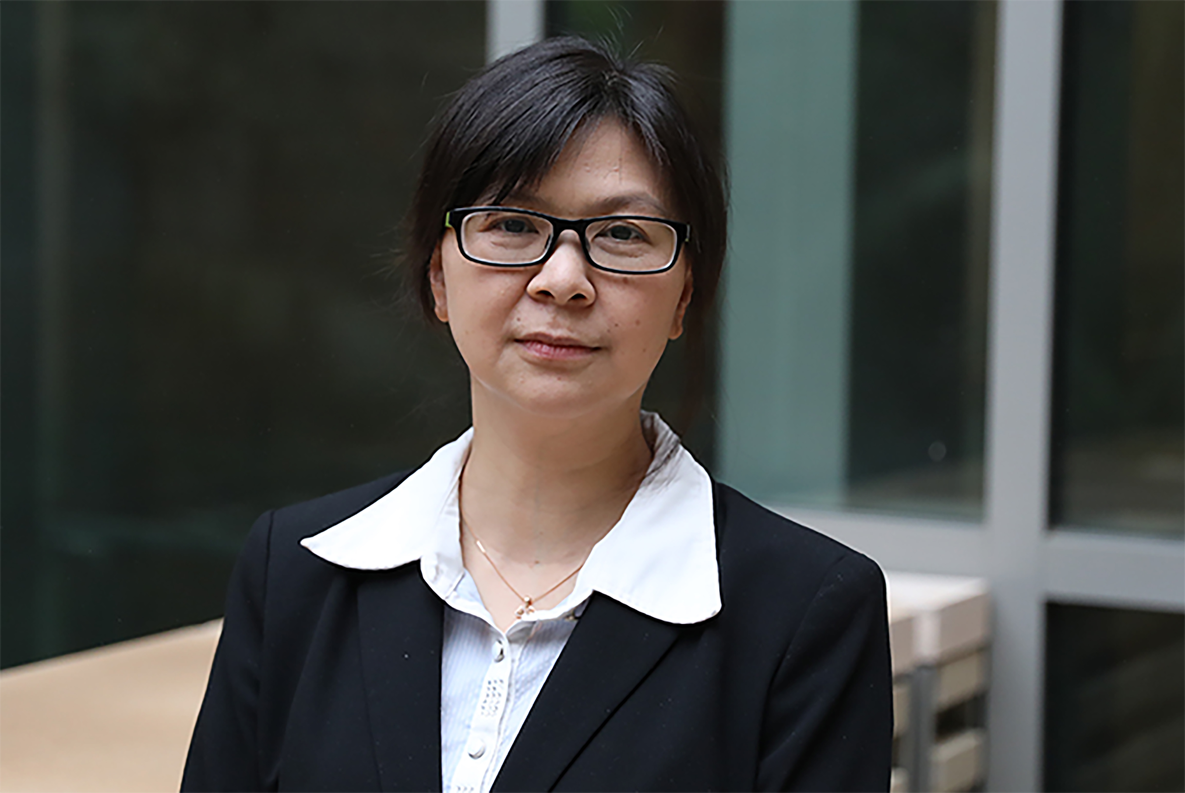Midrange shot of Yu Huang wearing a white blouse, dark jacket and glasses in front of a building.