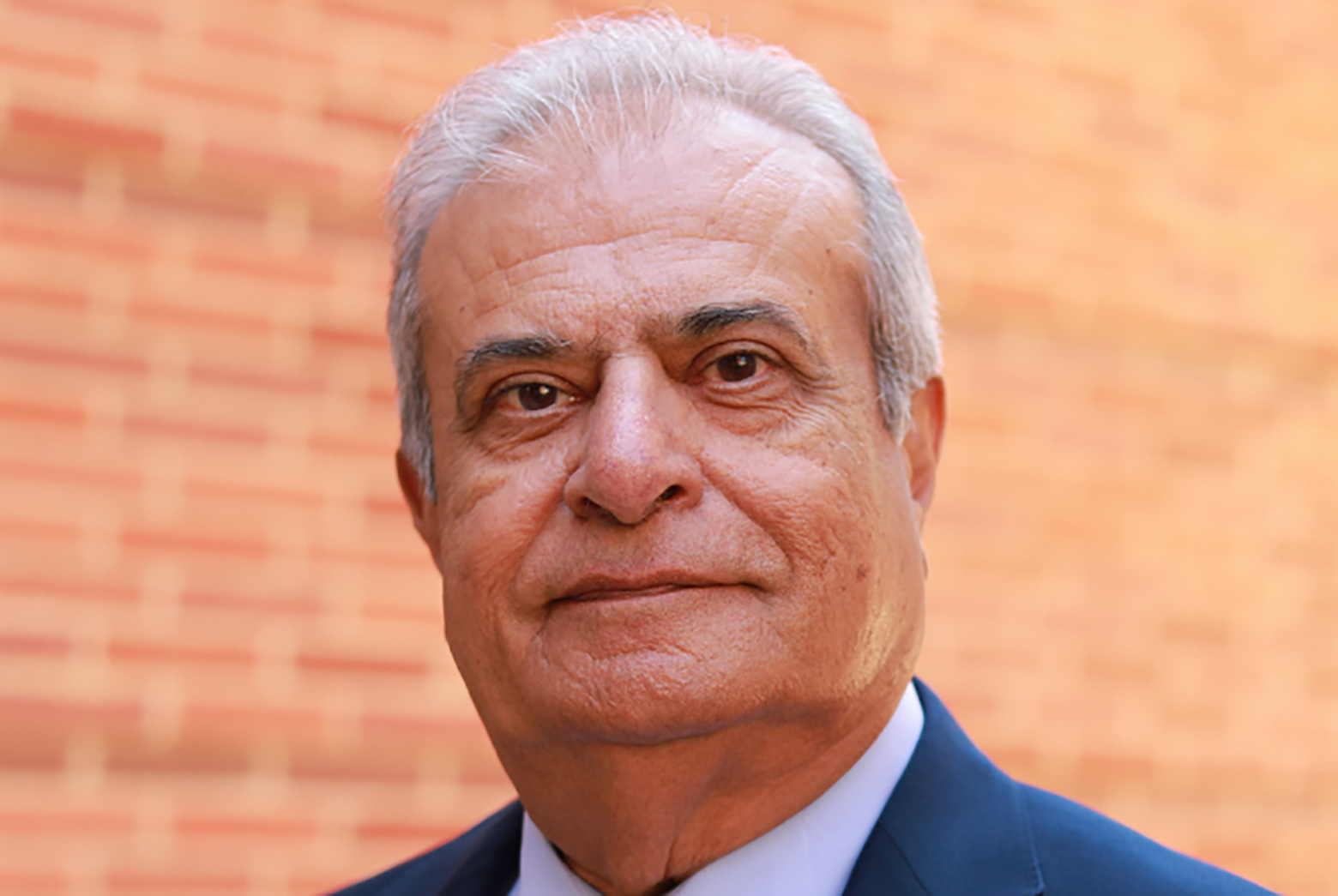Asad Madni wearing a navy suit and light blue shirt, in front of a blurred orange brick wall.