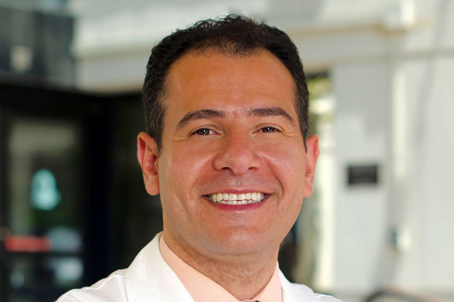 Close-up shot of Tamer Sallam wearing a white lab coat and pale orange shirt.