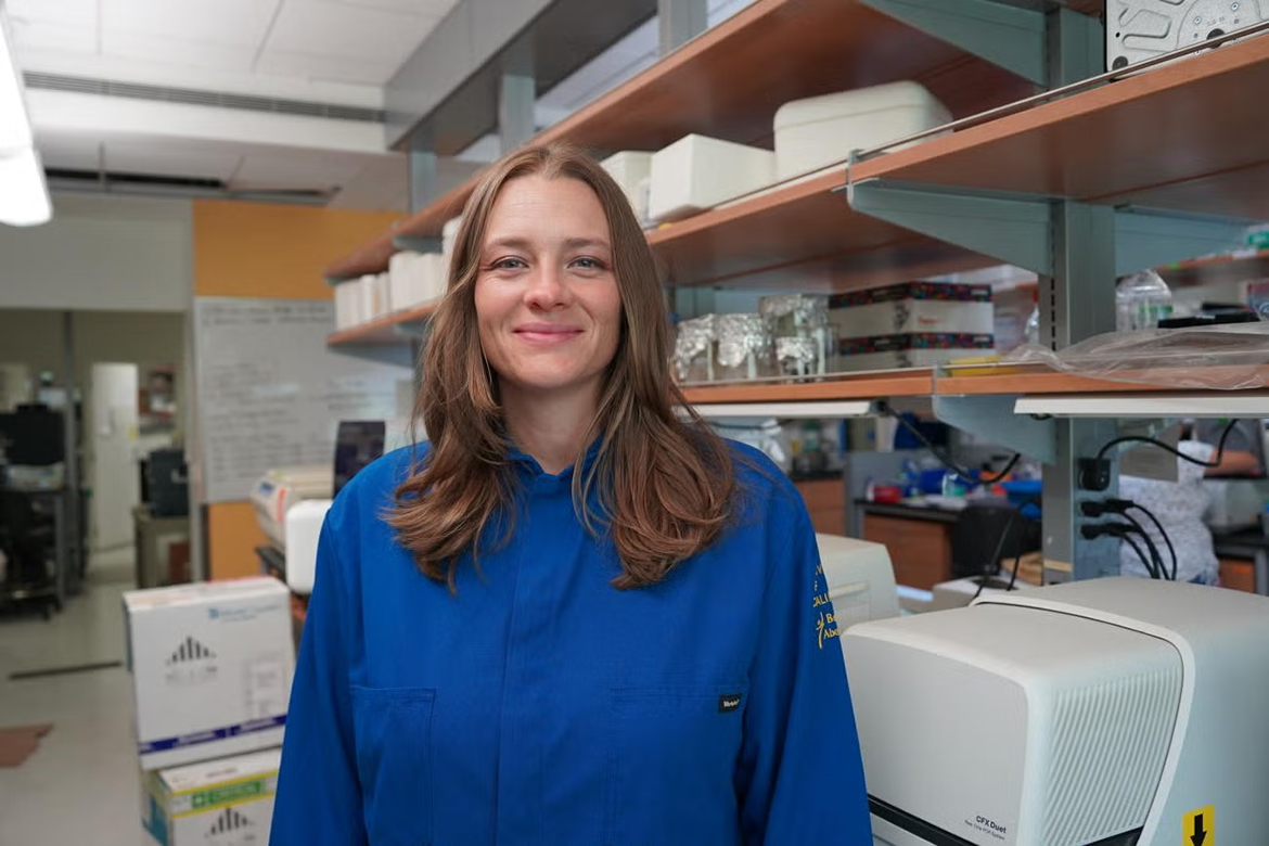 Tara TeSlaa, wearing a blue lab coat, stands in a laboratory surrounded by scientific equipment, storage shelves and research supplies.