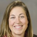 Jennifer DeVoe, M.D., has long light brown-blonde hair, a pink dress shirt and black blazer, smiling against a light brown background.
