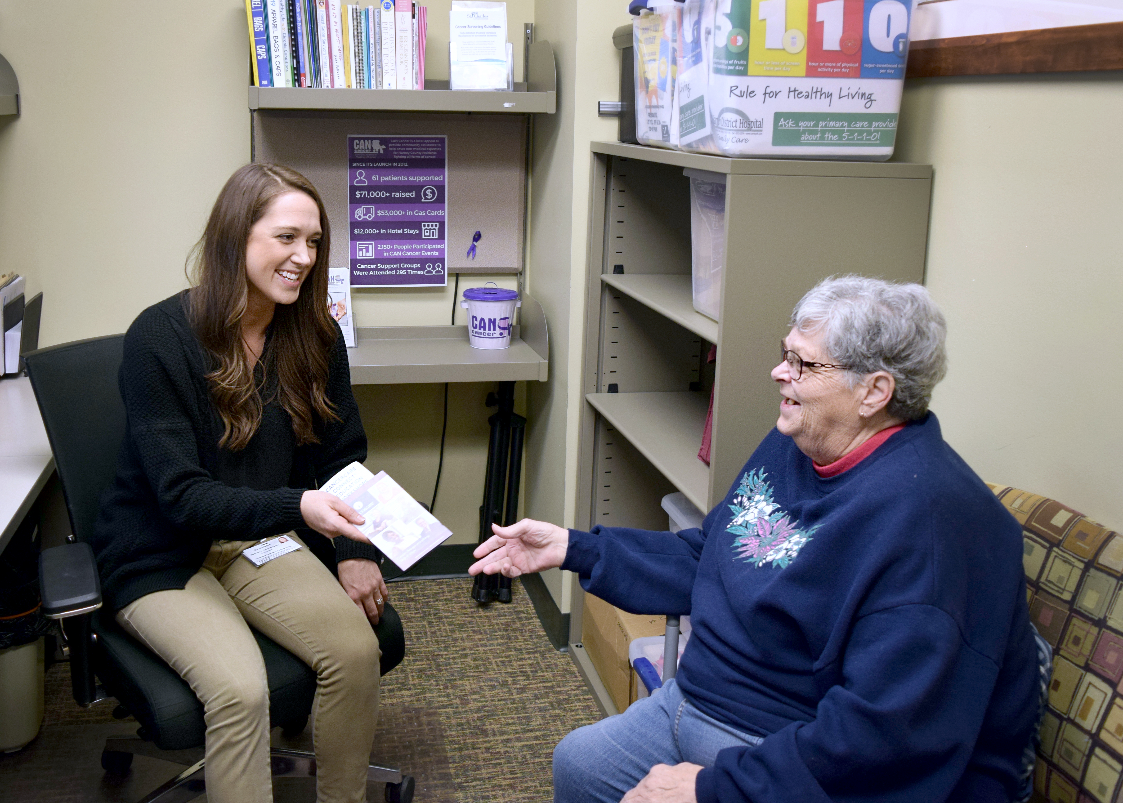 Savanna Cate, (left) program outreach coordinator for the Harney District Hospital Community Assistance for Neighbors with Cancer Program, shares information with patient Debbie Pike. (Photo courtesy Harney District Hospital Community Assistance for Neighbors with Cancer Program) two women sitting in an office, smiling at one another, seated.