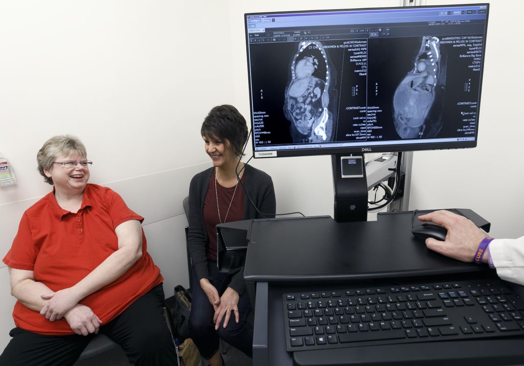 two women, seated and talking with another, with a computer monitor on the right side of the picture showing two xray scans, before and after, of the patient