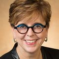 Nancy Haigwood (OHSU) has a black jacket, blue top, multicolor glasses and red-brown hair, smiling.