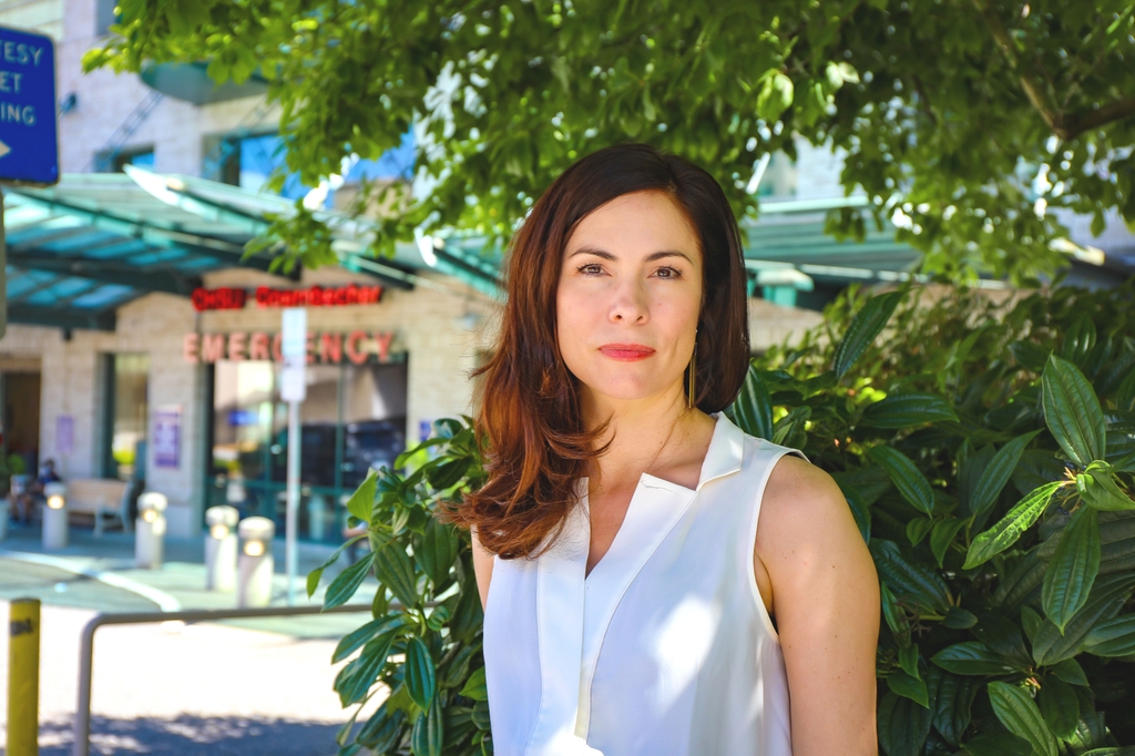 Kathleen Carlson, Ph.D., has long dark auburn hair, and is wearing a white top, standing near the OHSU Emergency Department entrance in background. 