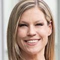 Abby Dotson, Ph.D. has long blonde hair, black top, standing in a hallway, smiling.