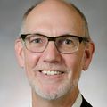 Jim Anderson, M.D., has a tan suit, short gray hair, eyeglasses, smiling against a gray background.