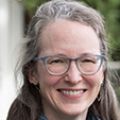 Moira Ray, M.D., M.P.H. has long gray/brown hair, glasses and an aqua-colored scarf, standing outside the BICC Library at OHSU.