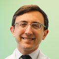 Ross Harrison, M.D., tiene el pelo corto y oscuro peinado hacia un lado, anteojos y usa una bata blanca en una habitación para pacientes en el centro de tratamiento de cáncer OHSU Knight.