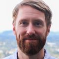 James Frank, Ph.D., has light reddish blond hair, a beard and a blue top, smiling with the Portland city scape behind him.