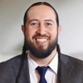 Claymore Kills First, Pharm. D., is wearing a suit, and has long dark hair into braids, and facial hair, smiling against a gray background.