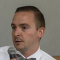 Bradley Buchheit, que tiene cabello castaño corto y viste una camisa blanca con una pajarita a cuadros, se sienta en la reunión del panel de información sobre fentanilo en OHSU.