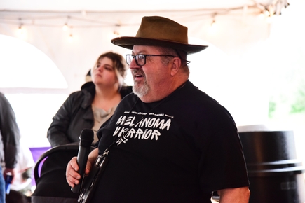 Robin Zimmerman (wearing a brimmed hat and eyeglasses, holds a microphone. Zimerman is a volunteer for Oregon’s Steps Against Melanoma Walks, OHSU Knight Cancer Institute science research advocate and panel member for the International Alliance for Cancer Early Detection, welcomes the crowd at Frog Pond Farms in Wilsonville on May 4, 2024. (OHSU/Patrick Kinghorn)