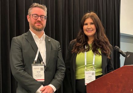 Aaron Kerr and Kayla Freudenthal, left to right. Aaron wearing a gray suit, and Kayla wearing a lime green top with black pants, on a stage at the ACMA conference.