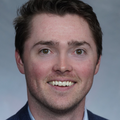 Dylan Robinson has short brown hair, a blue suit, and smiling against a gray background.