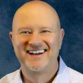 Mike Petitt is bald, wearing a light shirt and smiling against a blue background.