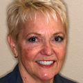 Ronda Trotman has short blonde hair, a dark top and smiling against a beige background.