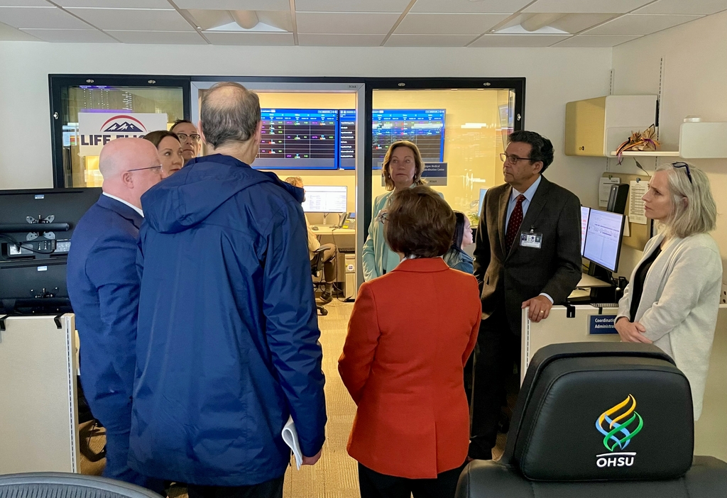 U.S. Senator Ron Wyden and U.S. Congresswoman Suzanne Bonamici tour the Oregon Behavioral Health Coordination Center within the OHSU Mission Control Center. Wyden and Bonamici encouraged further investment in coordinating resources and providing behavioral health services. image is of the backs of the senators dressed professionally facing the data screens at OBCC at OHSU and standing with a crowd of OHSU officials.